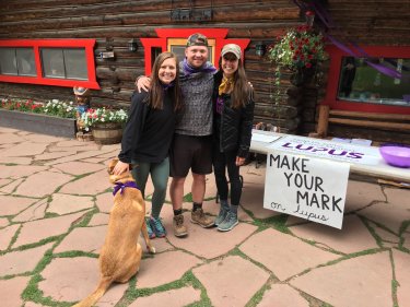 Three people stand next to a dog who is wearing a purple bandana. They are standing by a table that has a sign saying Make Your Mark on lupus.